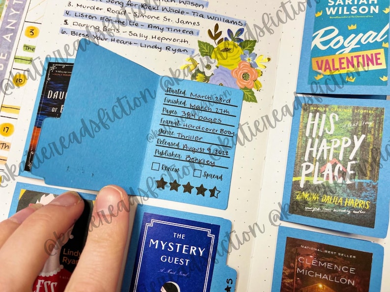 May include: A close-up of a blue book journal with a blue bookmark. The bookmark has a handwritten review of a book titled "The Mystery Guest". The review includes the date started, date finished, number of pages, format, genre, release date, publisher, and a 4-star rating.