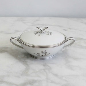May include: White porcelain sugar bowl with lid, featuring a delicate floral design in silver and green. The bowl has two handles and a silver trim around the rim.