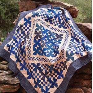 May include: A blue and white quilt with a "Blue Me Away" title and "Black Cat Creations" logo. The quilt is made with a variety of blue fabrics and has a white background. The quilt is folded and draped over a pile of rocks.