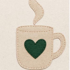 Könnte beinhalten: Eine Kaffeetasse aus beigem Filz mit einem grünen Herz und Dampf, der aus der Oberseite aufsteigt. Die Tasse hat einen Henkel und ist mit einem Kontrastfaden genäht. Der Hintergrund ist ein hellbeiger Stoff.