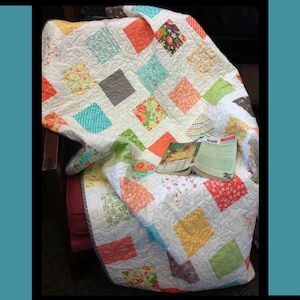 May include: A colorful quilt with a patchwork design in shades of pink, orange, yellow, blue, and green. The quilt is folded and draped over a chair. The quilt is titled "Salt Water Taffy" and is by Tricia Lynn Maloney, Orphan Quilt Designs for Villa Rosa Designs.