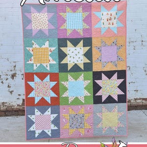 May include: A patchwork quilt with a star design, showcasing squares of patterned fabrics in pastel shades. The quilt is labelled "Amelia" and "Running Doe Quilts" for Villa Rosa Designs.