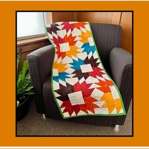 May include: A rectangular quilt featuring a star pattern in shades of orange, red, teal, and brown on a white background, finished with a bright green border. The text Shanagolden appears at the top, and Rachelle Handmade for Villa Rosa Designs at the bottom.