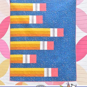 May include: A quilt pattern called "Pointless" featuring a blue background with yellow, orange, white, and pink pencil-shaped blocks. The quilt is made by Running Doe Quilts for Villa Rosa Designs.