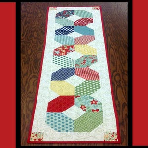 May include: A colorful table runner with a geometric pattern of hexagons in red, yellow, blue, green, and white. The runner is made of fabric and has a white background. The text "Penny Candy" is at the top of the image. The text "Tricia Lynn Maloney Orphan Quilt Designs For Villa Rosa Designs" is at the bottom of the image.