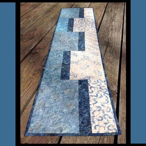May include: A quilted table runner with a blue and white ripple design. The runner features a patchwork pattern with various blue floral and patterned fabrics. The text "Ripple Runner" is at the top.