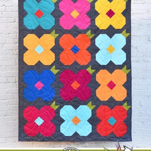 May include: A quilt pattern called "May Flowers" by Running Doe Quilts for Villa Rosa Designs. The quilt features a repeating pattern of colourful flowers with a dark grey background.