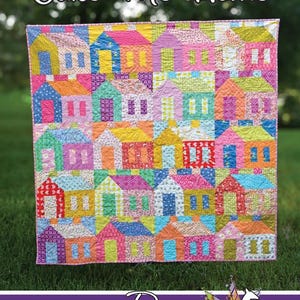 May include: A quilt featuring a grid of colorful house blocks in various patterns and bright colors. The text Cake Me Home is at the top, and the text Running Doe Quilts For Villa Rosa Designs with a deer graphic is at the bottom.