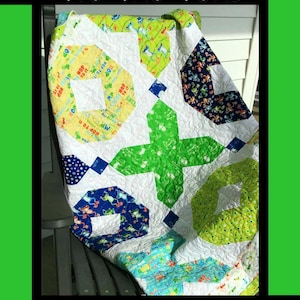 May include: A handmade quilt with a Tic Tac Toad design. The quilt features a white background with green, blue, and yellow geometric shapes. The quilt has the text "Tic Tac Toad" at the top and the designer's name and company at the bottom.