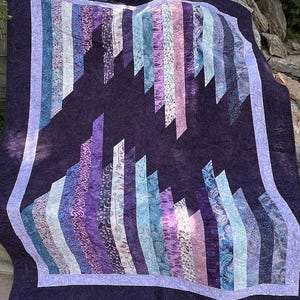 May include: A handmade quilt with a geometric design. The quilt features vertical stripes in shades of purple, blue, and white, set against a dark purple background. The quilt is bordered with a lighter purple fabric.