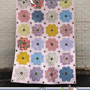 May include: A handmade quilt with a light pink background and a floral pattern. The quilt features a repeating design of colorful flower-shaped blocks. The quilt has the name "Maureen" at the top and the Running Doe Quilts logo at the bottom.