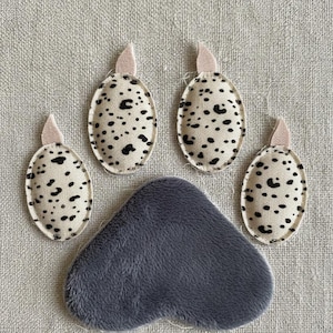 May include: A grey plush paw pad with four oval appliqués. The appliqués are cream-coloured with black spots, and each has a small pink accent. The items are arranged to resemble a dog's paw.