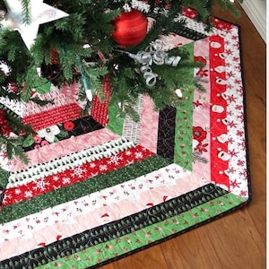 May include: A colorful Christmas tree skirt with a patchwork design. The skirt features red, green, pink, and black fabrics with snowflake, Santa, and floral patterns. The skirt is placed under a decorated Christmas tree with ornaments and lights.
