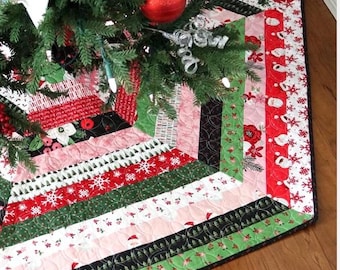 May include: A colorful Christmas tree skirt with a patchwork design. The skirt features red, green, pink, and black fabrics with snowflake, Santa, and floral patterns. The skirt is placed under a decorated Christmas tree with ornaments and lights.
