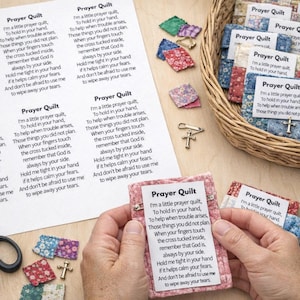 May include: A collection of small, handmade prayer quilts with printed text and a cross charm. The quilts are in a woven basket and held by a person. Additional fabric squares, scissors, and pins are also visible.