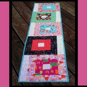 May include: A colorful, rectangular quilted table runner with a cupcake theme. The runner features various patterned squares in shades of blue, pink, and black, with the words "Cupcake Party" at the top. The design includes cupcake and kitchen-themed patterns.