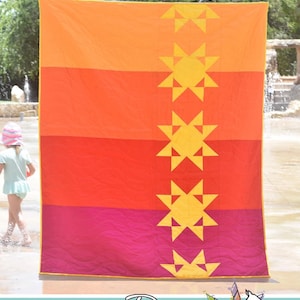 May include: Heat Wave quilt pattern by Running Doe Quilts for Villa Rosa Designs. The quilt features a vertical row of yellow stars on a background of orange, red, and pink fabric.