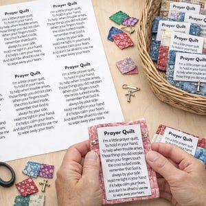 Puede incluir: Una colección de pequeños edredones de oración hechos a mano con texto impreso y un dije de cruz. Los edredones están en una cesta de mimbre y son sostenidos por una persona. También se ven cuadrados de tela adicionales, tijeras y alfileres.