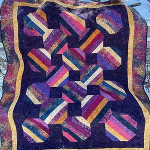 Könnte beinhalten: Eine handgefertigte Steppdecke mit einem dunkelvioletten Hintergrund und geometrischen Formen in verschiedenen Farben. Die Formen bestehen aus diagonalen Streifen in Rosa-, Orange-, Gelb- und Lilatönen. Die Steppdecke hat einen goldenen Rand.