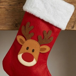 Puede incluir: Calcetín navideño rojo con puño de piel sintética blanca. El calcetín presenta una aplicación de cara de reno con cuernos marrones, cara beige y nariz roja. Se adjunta un lazo rojo para colgar. Decoración festiva navideña.