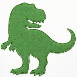 Può includere: Un'applicazione in tessuto verde a forma di dinosauro Tyrannosaurus Rex. La sagoma presenta una superficie testurizzata con cuciture visibili lungo tutto il bordo esterno, che definiscono la testa, le braccia corte, le zampe e una lunga coda curva.