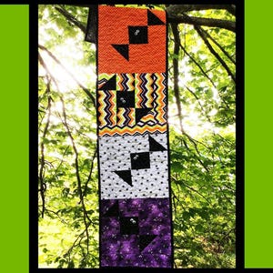 Puede incluir: Un edredón con temática de Halloween con el texto "Bat Dance" en la parte superior. El edredón presenta secciones naranjas, verdes, blancas y moradas con patrones de murciélagos y zig-zag. El edredón se exhibe al aire libre, colgado de una rama de árbol.