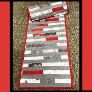 Op de afbeelding: Een handgemaakte quilt met de tekst "Sew Random". De quilt heeft een grijze achtergrond met horizontale strepen van rode, witte en bedrukte stof. De quilt is rood omrand.