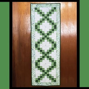 May include: A green and white table runner with a shamrock design. The table runner is called "Erin" and is by Tricia Lynn Maloney for Villa Rosa Designs.