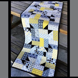 May include: A quilted table runner with a patchwork design. The runner features a mix of yellow, blue, gray, and black geometric patterns. The word "Dapper" is at the top. The text "Tricia Lynn Maloney, Orphan Quilt Designs, For Villa Rosa Designs" is at the bottom.