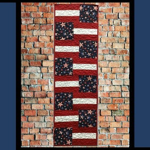 May include: A patriotic quilt design against a brick wall. The quilt has red and white stripes with navy blue squares filled with white stars. The word "Firecracker" is at the top.