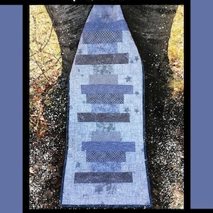 May include: A quilt with a blue and gray color scheme, featuring the text "Frosted Window Panes." The quilt has a rectangular shape with horizontal stripes of varying patterns and shades of blue and gray. The design is set against a backdrop of a tree trunk.