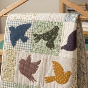 Può includere: Una trapunta patchwork con applicazioni di uccelli in vari colori e motivi, tra cui quadretti blu, stampe floreali e tessuti a tinta unita. Gli uccelli sono cuciti sulla trapunta, appesa a un appendiabiti di legno.