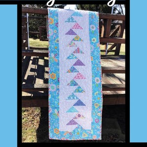 Op de afbeelding: Een gewatteerde tafelloper met een wit midden met rijen kleurrijke driehoekvormen. De loper is omrand met een blauw bloemenpatroon en de tekst "Fly Away". Het ontwerp is van Tricia Lynn Maloney.