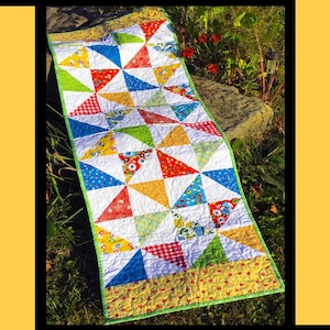 May include: A colorful quilt with a pinwheel design, featuring triangles in red, yellow, blue, and green. The quilt has a white background and a green border. The text "Pinwheel Picnic" is at the top.
