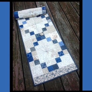 May include: A quilted table runner with a floral design and the text "Blueberry Cobbler." The runner features a patchwork design with blue, white, and gray squares. The top section is rolled up. The bottom border has a floral pattern.