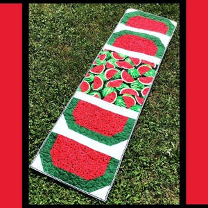 May include: A watermelon-themed table runner with a green and white background. The runner features a repeating pattern of watermelon slices. The text "Slice of Summer" is at the top of the image. The text "Tricia Lynn Maloney Orphan Quilt Designs For Villa Rosa Designs" is at the bottom of the image.