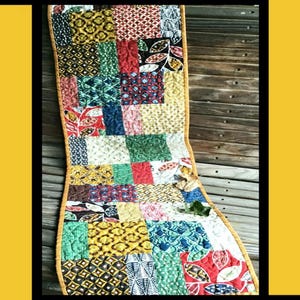 May include: A colorful patchwork quilt runner with a yellow border. The runner is made of various fabrics with geometric and floral patterns. The quilt runner is displayed on a wooden surface. The text "MOD TILES" is at the top of the image. The text "Tricia Lynn Maloney Orphan Quilt Designs For Villa Rosa Designs" is at the bottom of the image.