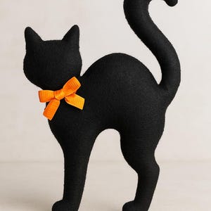 以下が含まれることがあります： オレンジ色のリボンが付いた黒猫の装飾品。猫は曲がった尻尾をして立っています。このアイテムはおそらくフェルトまたは同様の生地で作られており、ハロウィーンまたは季節の装飾です。