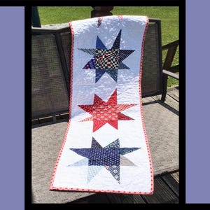 May include: A white Crazy Stars quilt with three large star patterns in red and blue patchwork. The quilt has a red and white border and is displayed on a metal chair. The text "Crazy Stars" is at the top.