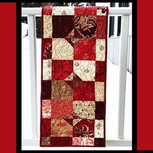 May include: A patchwork table runner in red, cream, and brown hues, displayed on a white railing. The runner features a variety of patterned fabrics. The text "Ruby Road" is at the top, with "Tricia Lynn Maloney" and "Orphan Quilt Designs" at the bottom.