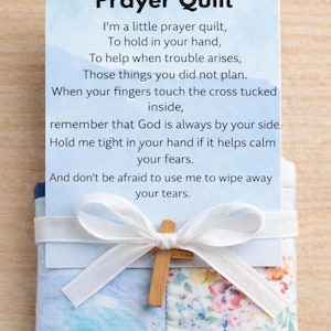 Puede incluir: Un edredón de oración con una cruz de madera y una cinta blanca. El edredón tiene un patrón azul y floral. El texto en la tarjeta dice "Prayer Quilt" y un poema sobre consuelo y fe.