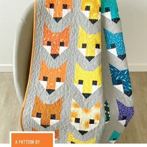 May include: A gray quilt featuring a repeating pattern of stylized fox faces in various colors, including orange, yellow, and teal. The quilt is folded over a chair. The text "FANCY FOX" is at the top.