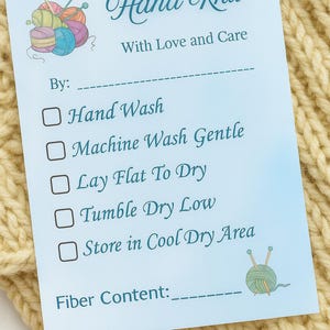 Könnte beinhalten: Eine hellblaue Pflegehinweiskarte mit den Worten "Hand Knit" und "With Love and Care". Die Karte enthält Waschanweisungen und einen Platz für den Fasergehalt. Die Karte liegt auf einem cremefarbenen Strickstoff.