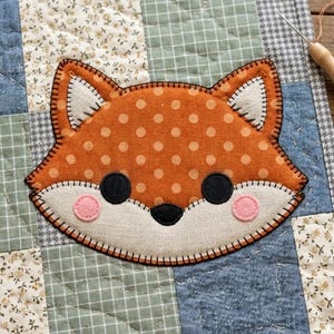 May include: A handmade fox applique on a patchwork quilt. The fox has orange polka-dot fur, black eyes, and pink cheeks. The quilt features various fabric patterns in blue, green, and floral designs, with a gray border.
