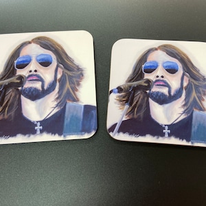May include: Two square coasters with a painting of a man with long brown hair, a beard, and sunglasses singing into a microphone. The man is wearing a black shirt with a silver cross necklace. The coasters are white with a light brown border.