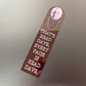 May include: A black metal bookmark with a white image of a man's face and the text "That's read Dave, every page is read Dave."