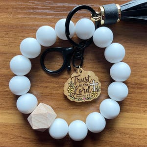 May include: A circular keychain bracelet featuring matte white silicone beads, one faceted natural wood bead, and a black leather tassel. A wooden charm is attached, engraved with the text Trust in the Lord Proverbs 3:5 and a cross.