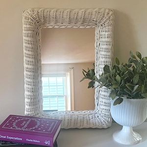 May include: A rectangular mirror with a white woven wicker frame. Beside the mirror sits a white fluted pedestal vase containing green foliage, and a stack of two books titled Great Expectations and Oliver Twist by Charles Dickens.