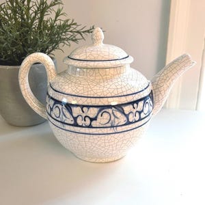 May include: A white ceramic teapot with a crackle glaze and blue decorative band. The teapot has a curved spout, a rounded body, and a lid with a small knob. A green plant in a gray pot is in the background.