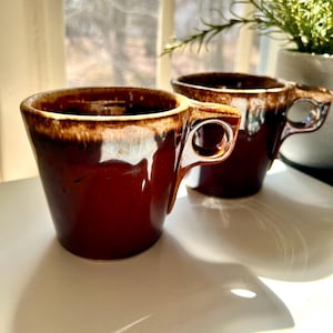May include: Two brown ceramic mugs with a unique handle design. The mugs have a glossy finish with a darker brown rim and a lighter brown interior. They are placed on a white surface near a window, with a plant in the background.